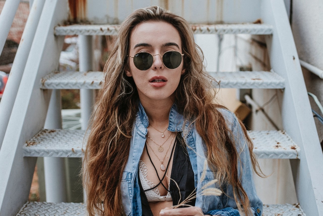 A woman sitting at the end of stairs with multiple necklaces layered under her multiple layers of bathing suit, shirt, and jacket