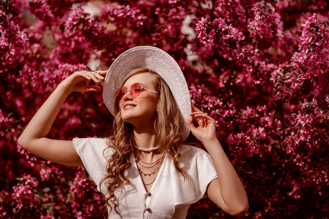 Wearing a white dress, rose colored sunglasses, and white hat with multiple necklaces that match her outfit as she stands in front of flowers