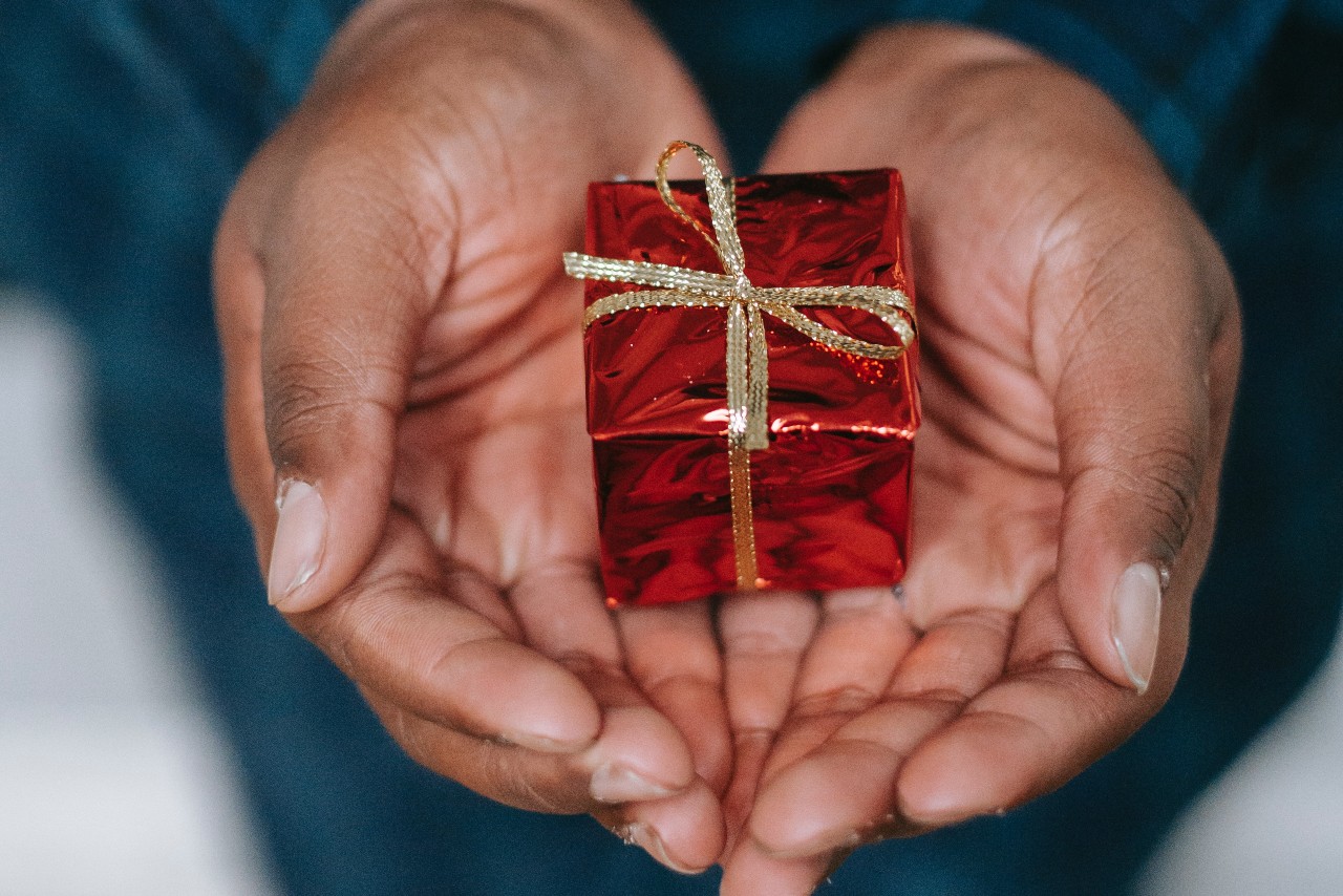 A person holding a present wrapped in shiny red paper along with a gold ribbon tied around it as well