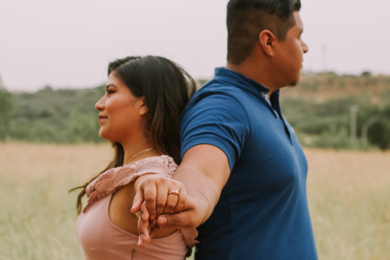 An engaged couple stands back to back and holds hands