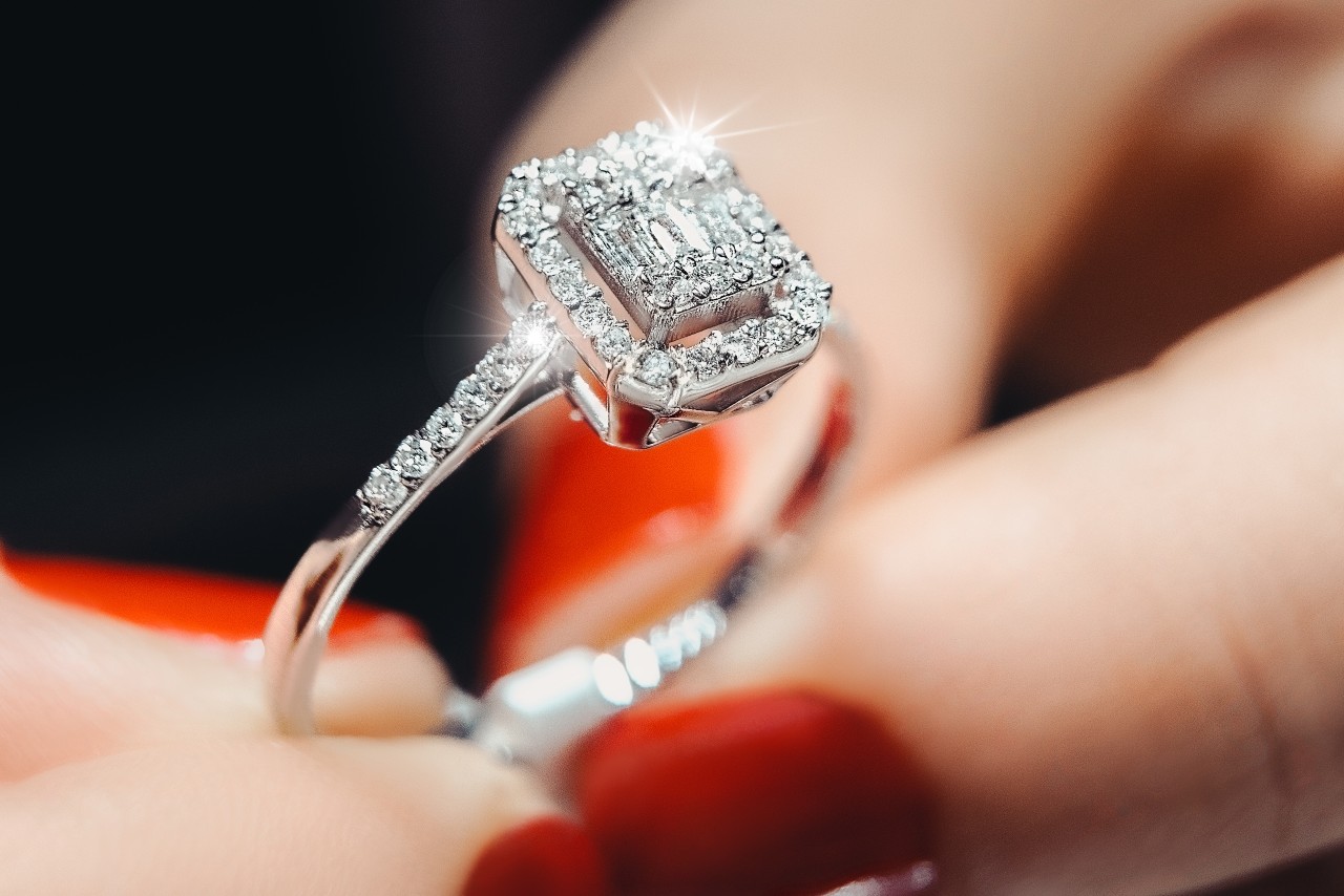 Close up image of a person with red painted nails holding a white gold, halo engagement ring