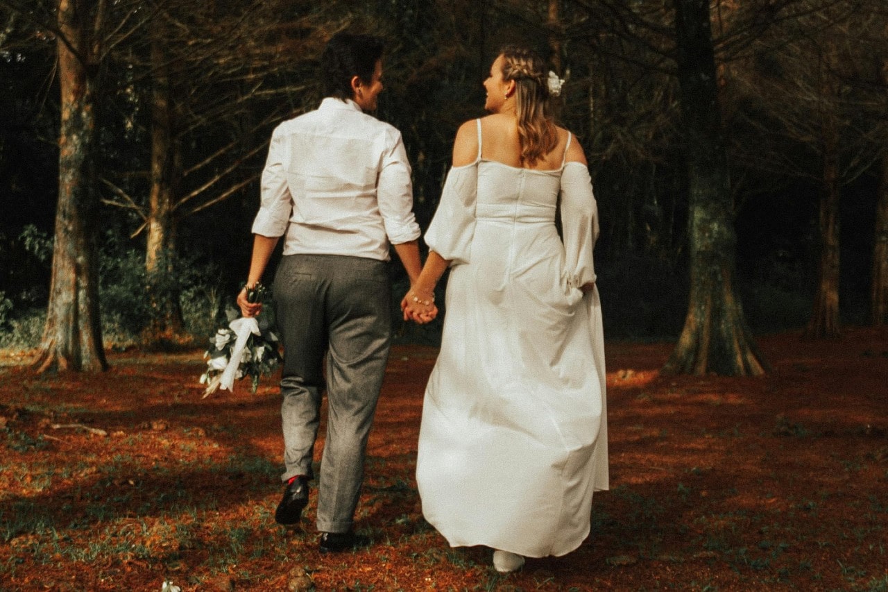 A couple walks hand-in-hand through a forest, one in a white gown, the other in a white shirt and gray pants holding a beautiful bouquet of white roses.