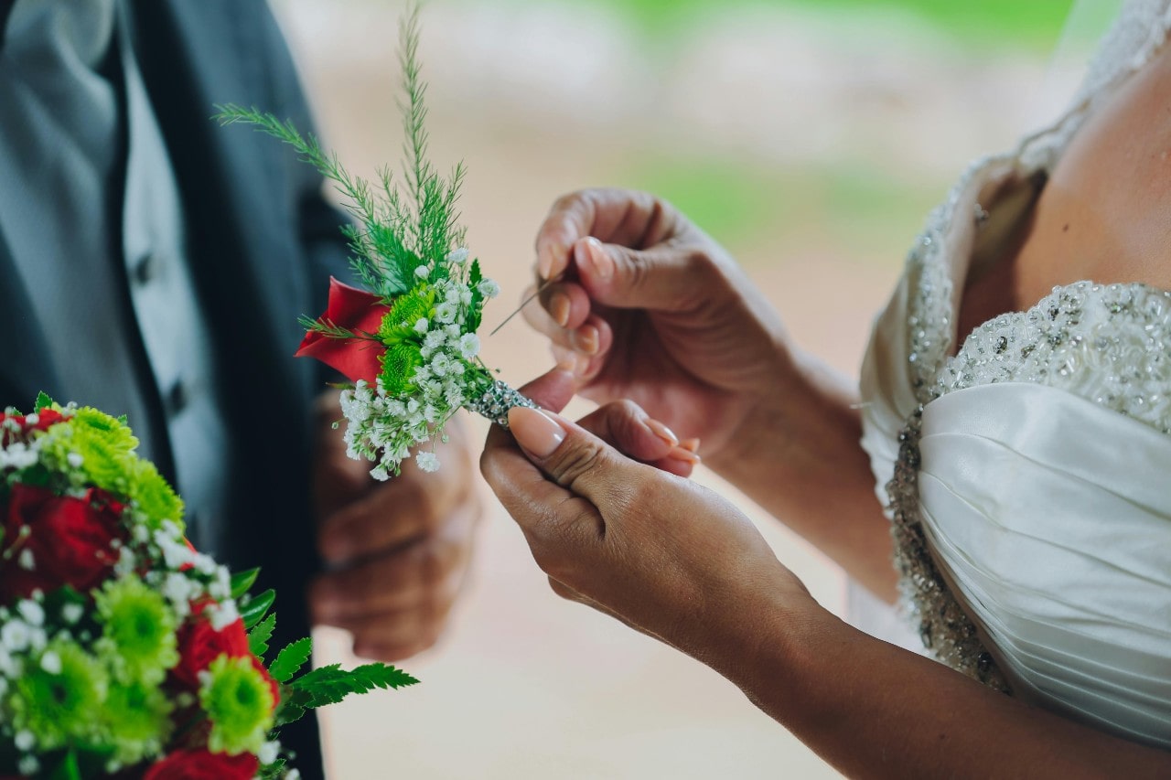 A bride in a white beaded gown pins a boutonniere with red and white flowers on a groom in a dark suit, against a blurred green background.