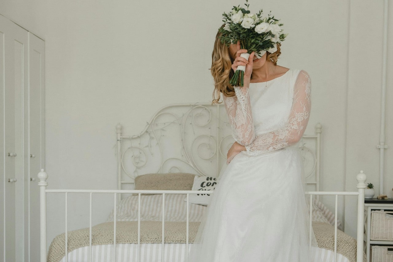 The bride in a white lace wedding dress sits on a bed, playfully covering her face with a bouquet of white flowers.