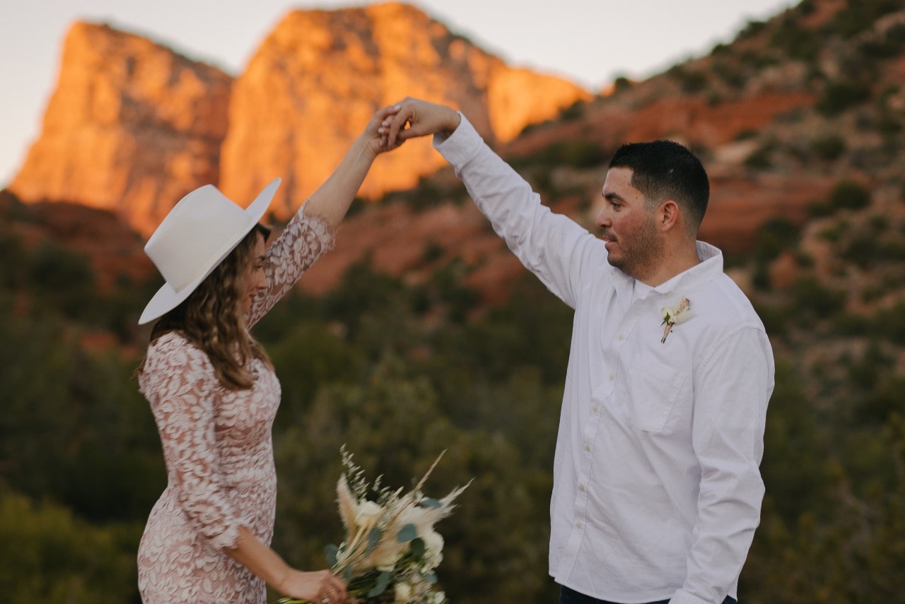 A couple dances joyfully in front of red rock formations at sunset. The woman wears a lace dress and white hat, holding flowers, while the man wears a white shirt.