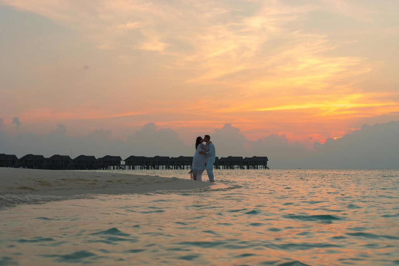 A couple kissing with their feet dipped into the ocean water, against a beautiful sunset.