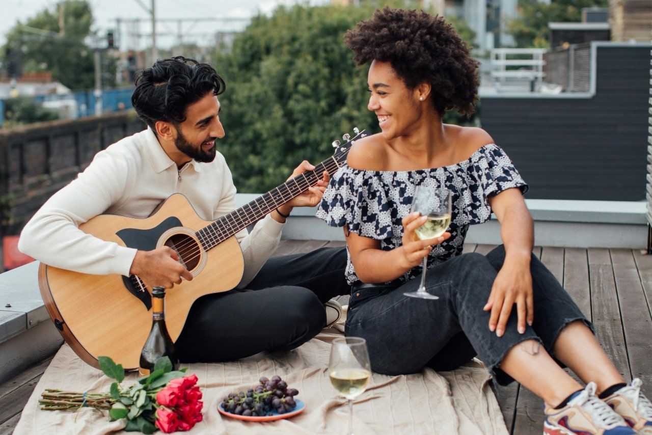 A couple enjoys a rooftop date; a man strums a guitar while a woman smiles, holding a glass of white wine. Red roses and grapes rest nearby.