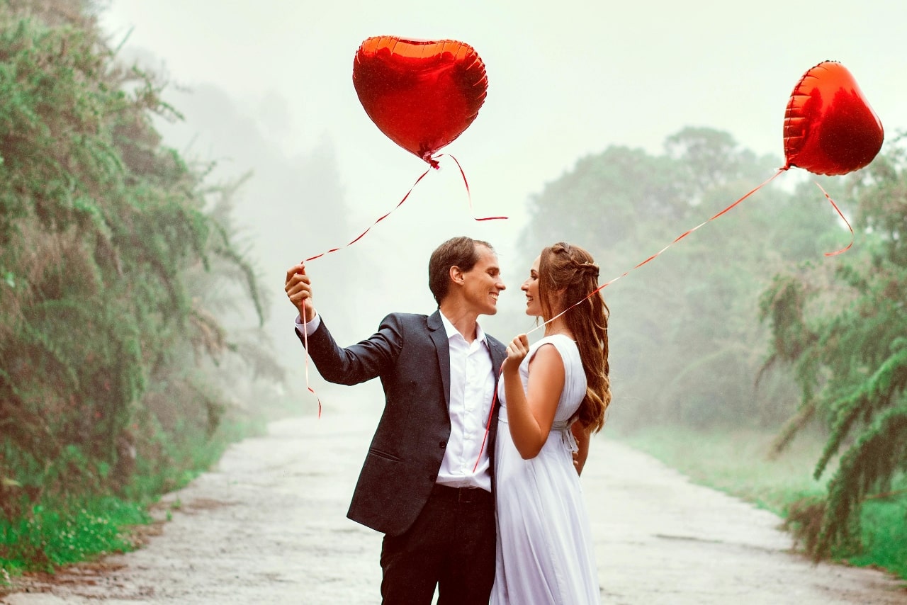 A couple stands on a misty road, smiling at each other, holding heart-shaped red balloons.