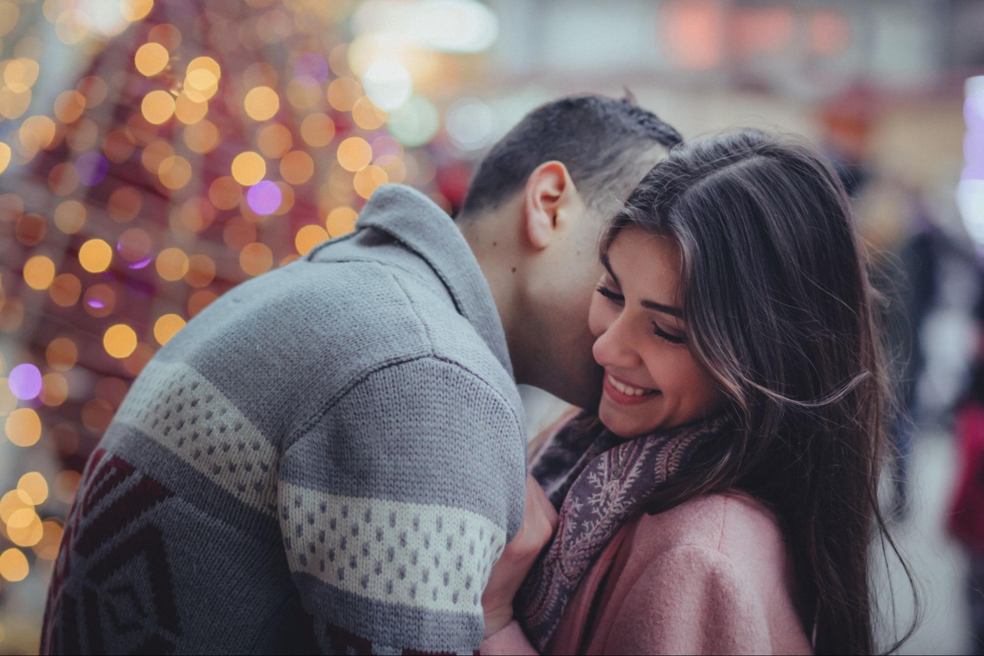 A couple shares a tender moment in front of a lit Christmas tree, surrounded by soft bokeh lights.