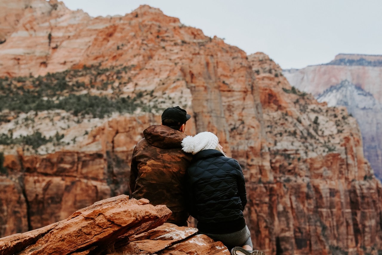 A couple, dressed warmly in jackets and a hat, sits closely together on a rocky ledge, overlooking vast, rugged red canyon cliffs under an overcast sky.