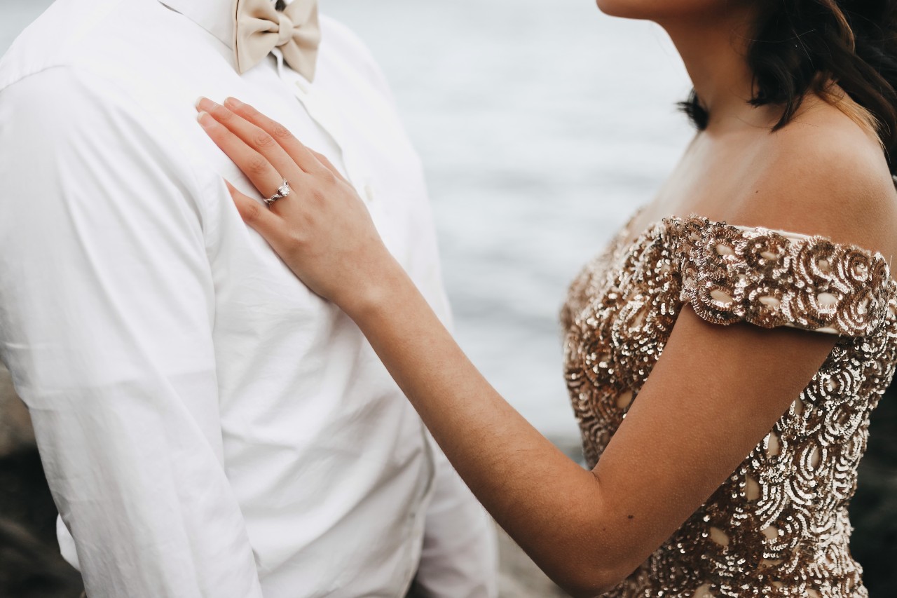 A bride in a glittering yellow gown puts a hand on a groom in a white shirt against an ocean view.