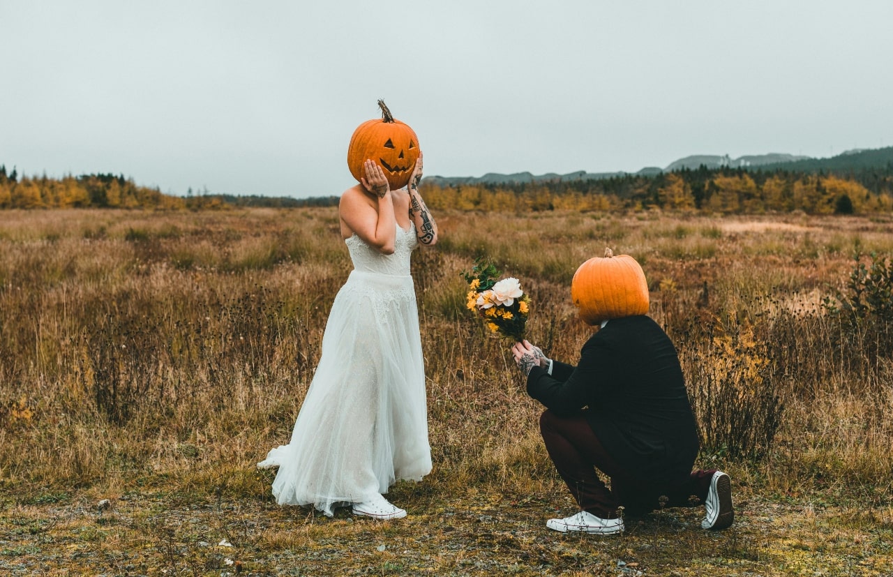 Bride and groom with pumpkins on heads in an autumn-ish outdoor backdrop.