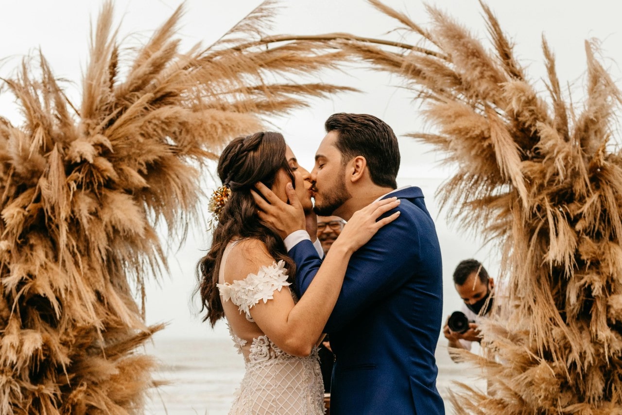 A newlywed couple kisses in front of the decorative arc, while the photographer takes a picture in the background.