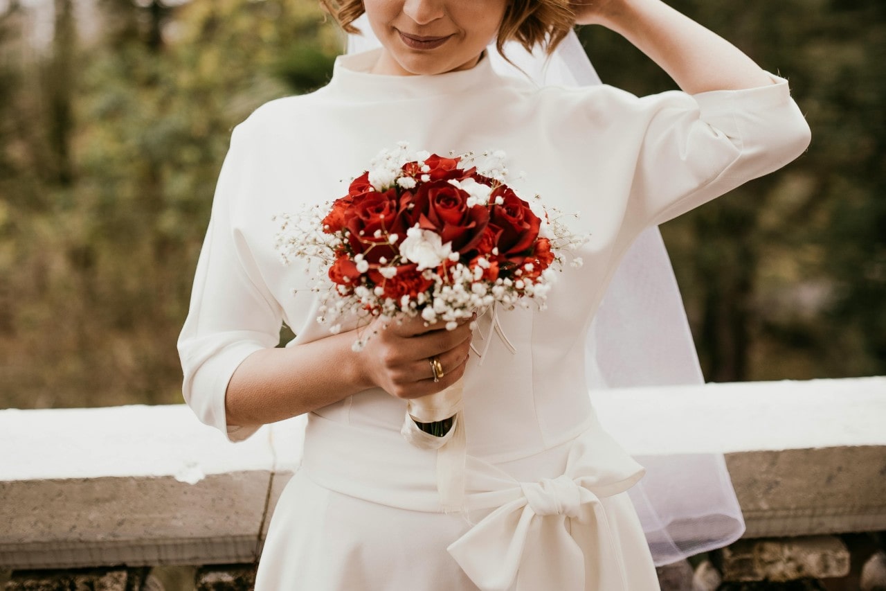 A bride in a ¾ sleeve wedding gown holds a bouquet of red roses in front of a blurred forestry background.