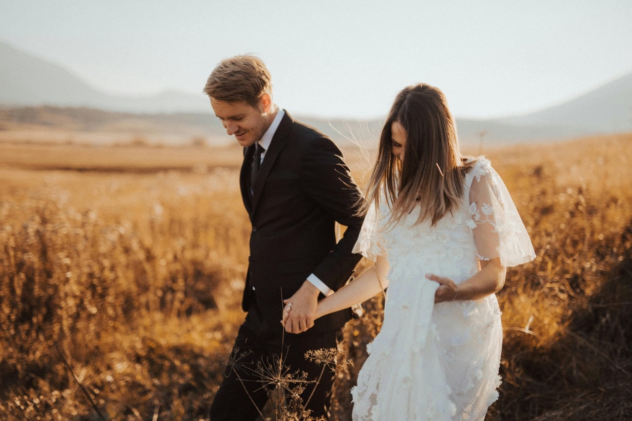A newlywed couple walk together hand in hand in a sunlight field full of wheat.