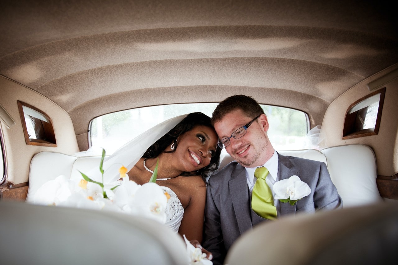 A newlywed couple sits in the back of the car smiling and leaning into each other.