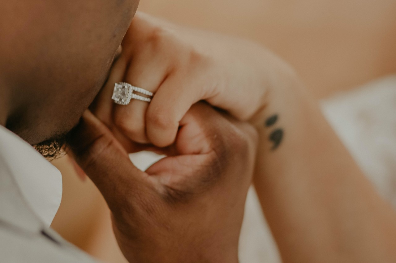 A man kisses the hand of his partner, wearing a halo engagement ring and wedding band.