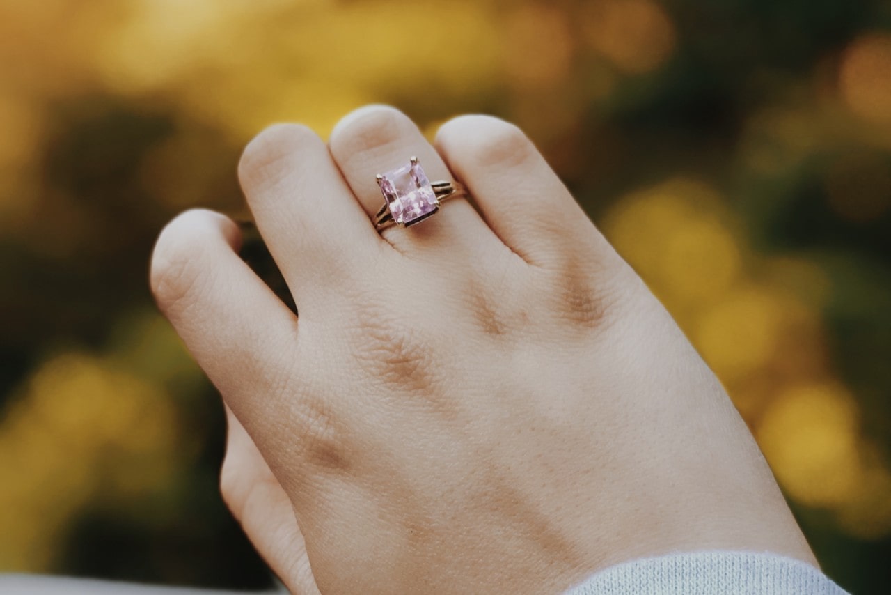 A close-up of a woman’s hand wearing a gold ring set with a large square-cut pink gemstone against a blurred natural background.
