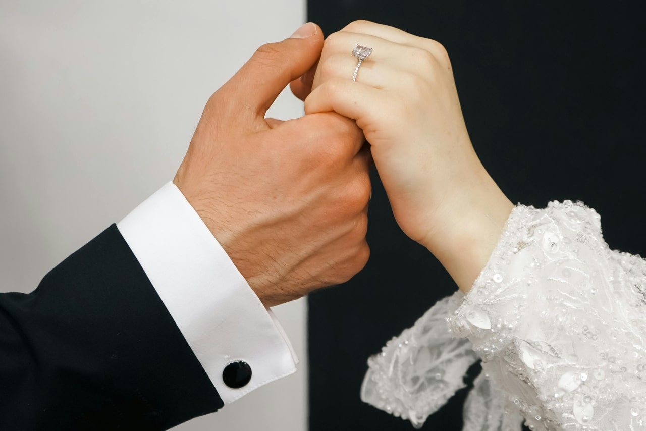 A bride and groom holding hands with a black and white background with a focus on the white gold diamond side stone engagement rings.