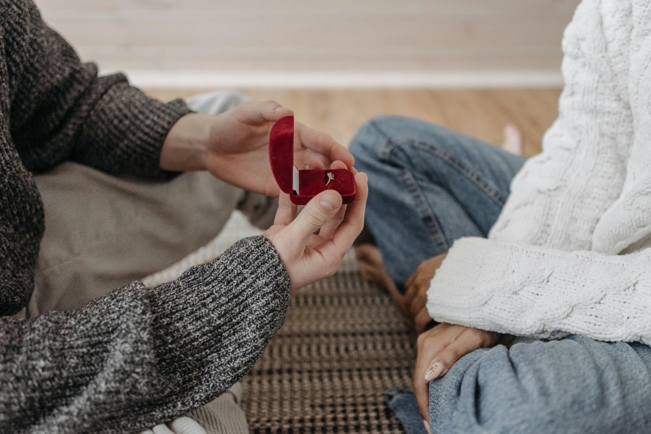 A person holding a red ring box and proposing while they sit across from each other on the floor