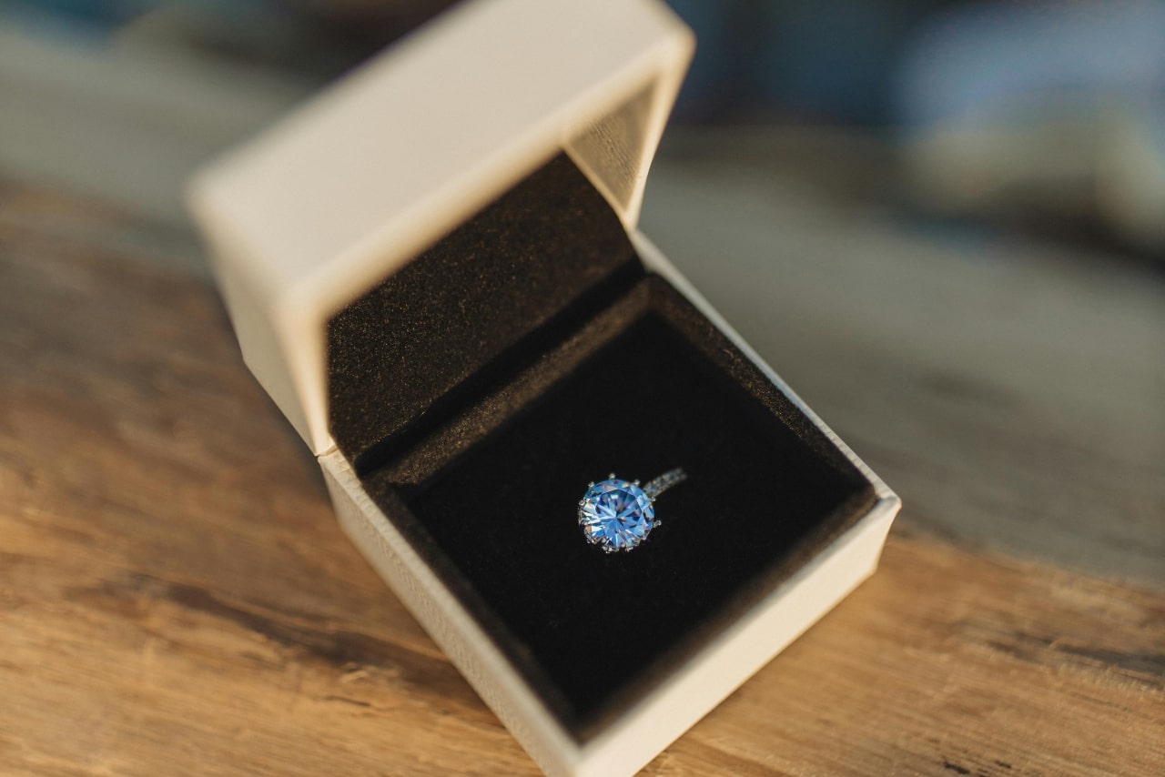 An elegant engagement ring with a large, sparkling diamond side stone ring in an open beige velvet jewelry box, resting on a wooden surface.