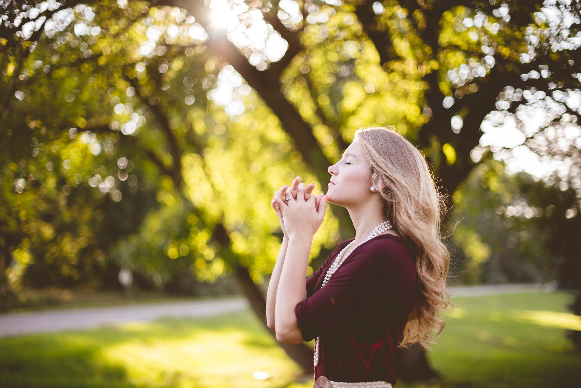 A young woman in a burgundy top stands in a sunlit park, eyes closed and hands clasped near her face, wearing a layered pearl necklace and a sparkling diamond ring, bathed in soft golden light through the trees.