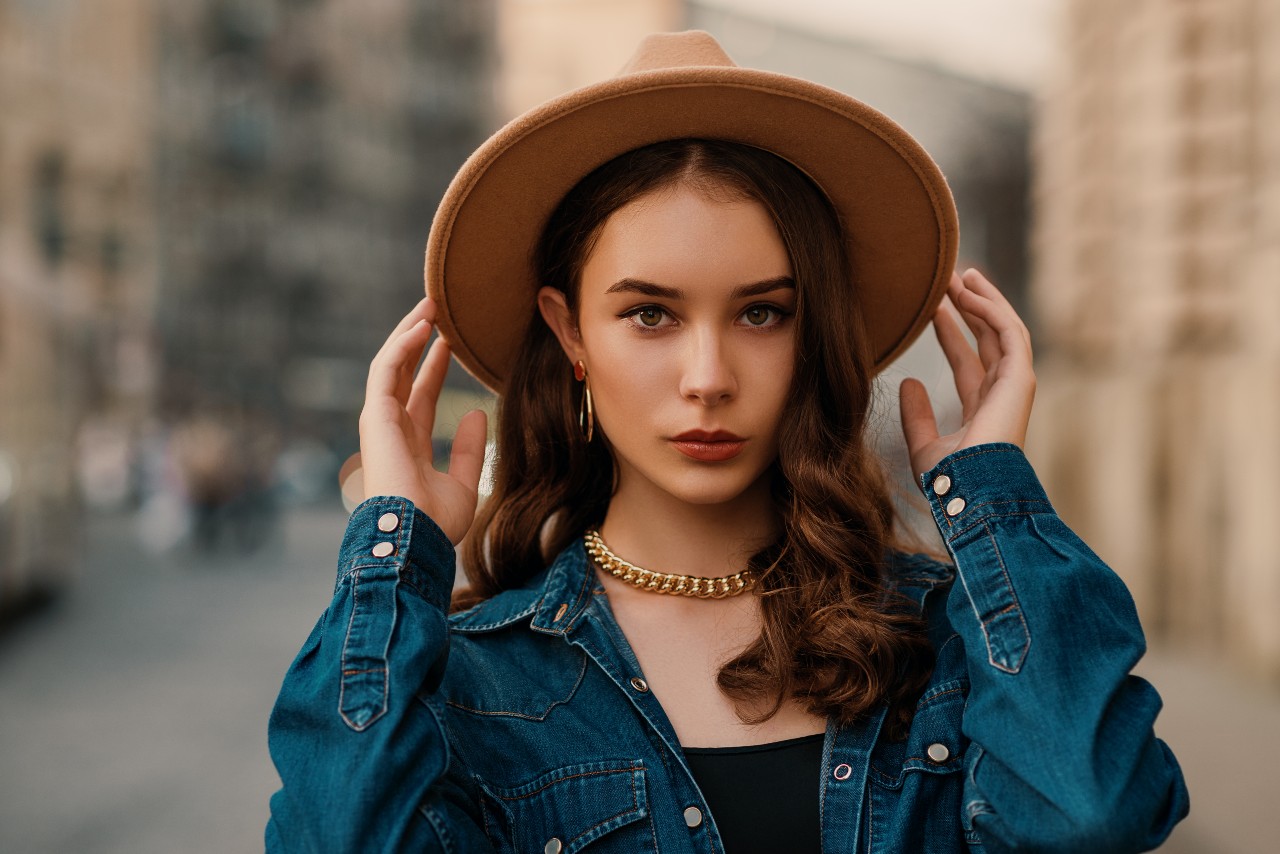 A young woman in a denim shirt and brown hat wearing a chunky gold chain necklace and hoop earrings