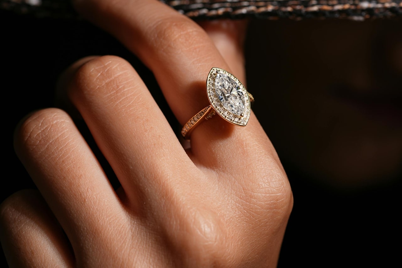 A close-up of a hand wearing an elegant gold ring with a large, marquise-cut diamond, set against a dark background.