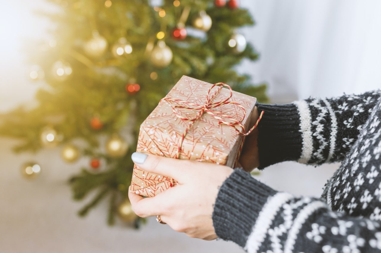 A close-up of a person in a black and white holiday sweater holding a festive gift box in front of a decorated Christmas tree.
