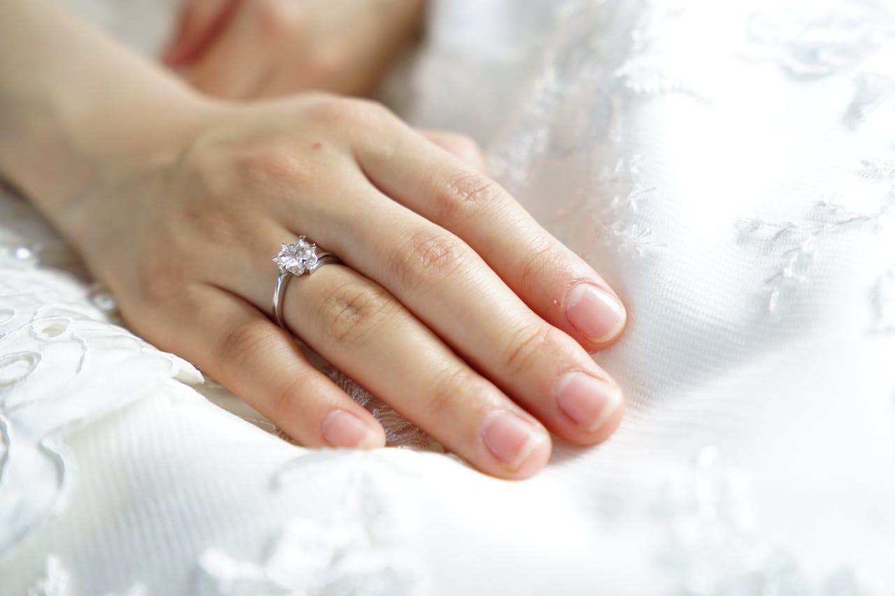 A close-up of a woman’s hand resting on an elegant wedding dress, with a delicate solitaire engagement ring on her finger.