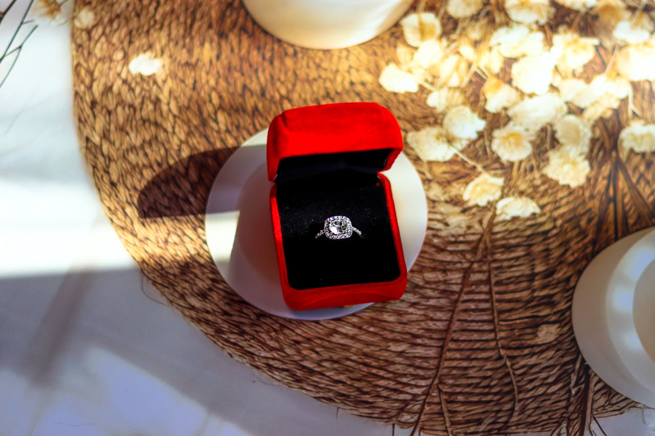 An overhead view of an elegant engagement ring displayed in-box atop a wicker pad with dried flowers.