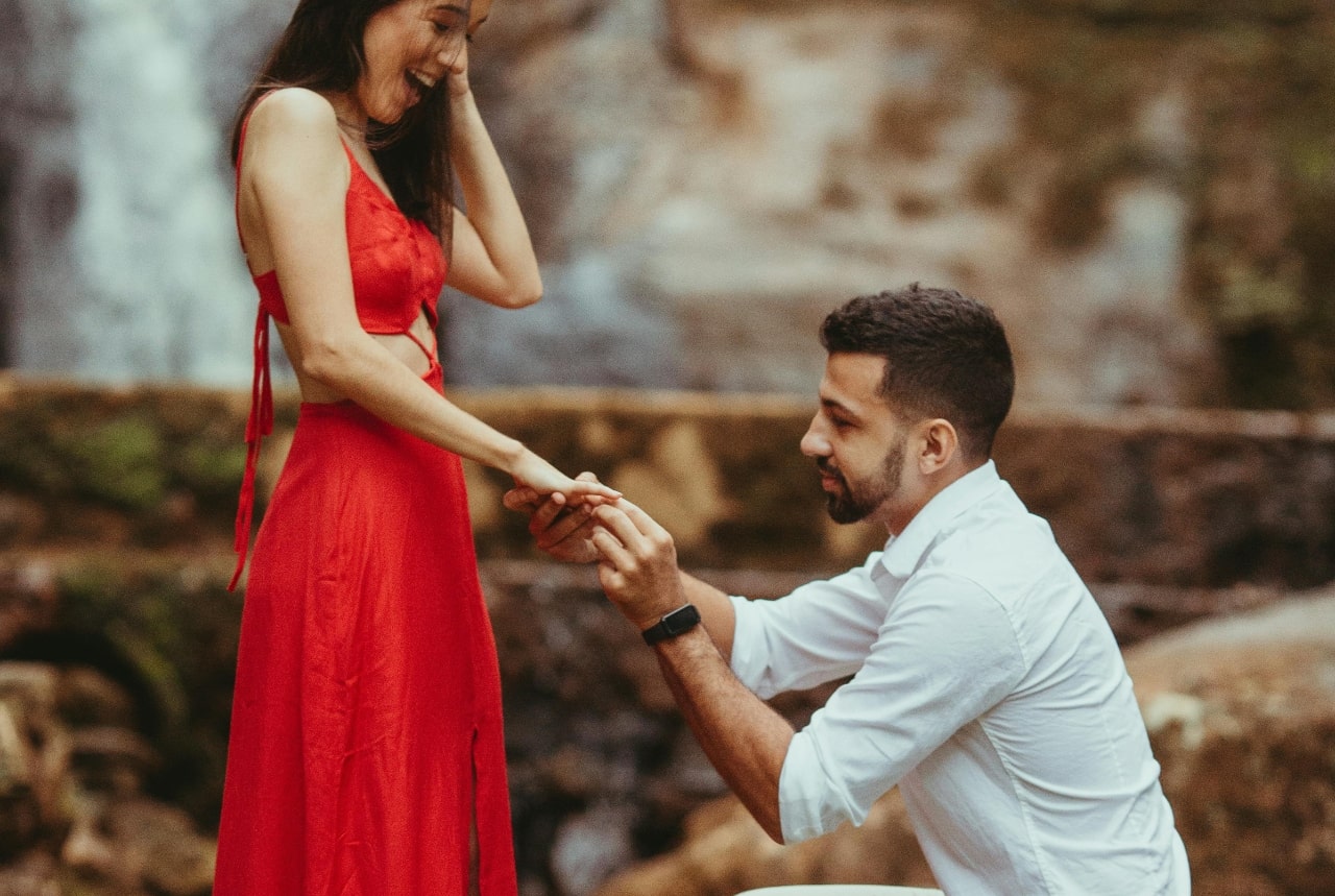 A close-up of a young man putting an engagement ring on his bride-to-be’s finger within an amphitheatre.