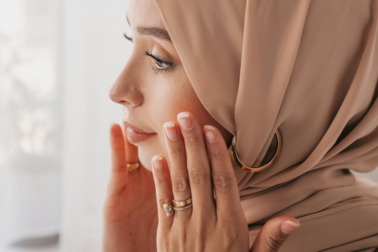A close-up of a woman wearing a tan hijab, with a glistening bridal stack on her finger.