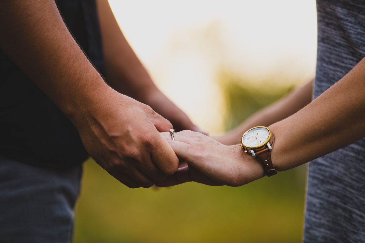 A couple holds hands outdoors, the woman wearing a white gold solitaire engagement ring and a watch.