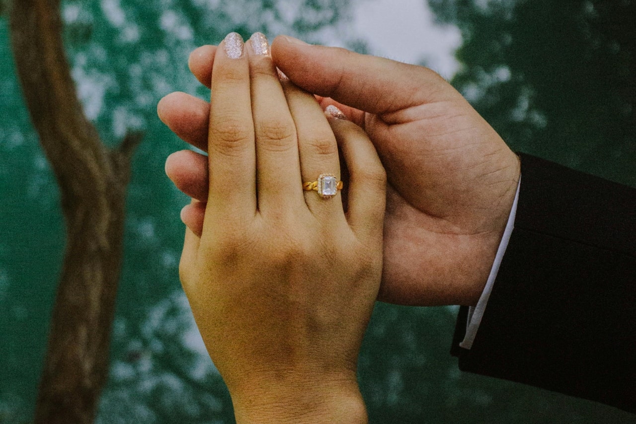 A couple holding hands in the forest showing her yellow gold, emerald cut diamond with round cut diamonds surrounding the center stone