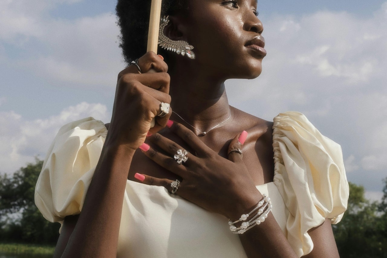 A woman wearing a cream puff-sleeved dress holding her hand to her chest and showing off her many silver jewelry pieces, standing in front of a field.