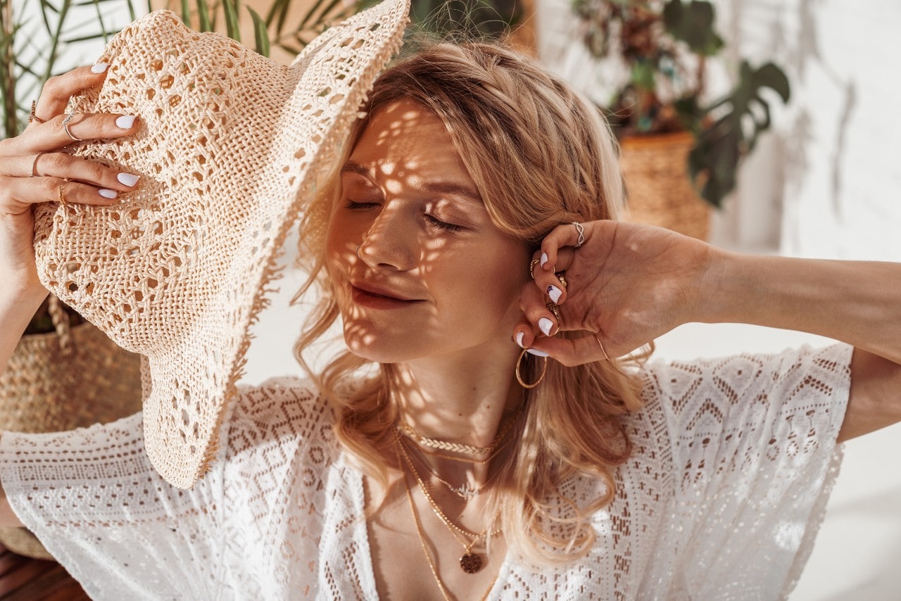 A woman shading herself with a straw hat while she wears multiple pieces of yellow gold jewelry A woman shading herself with a straw hat while she wears multiple pieces of yellow gold jewelry