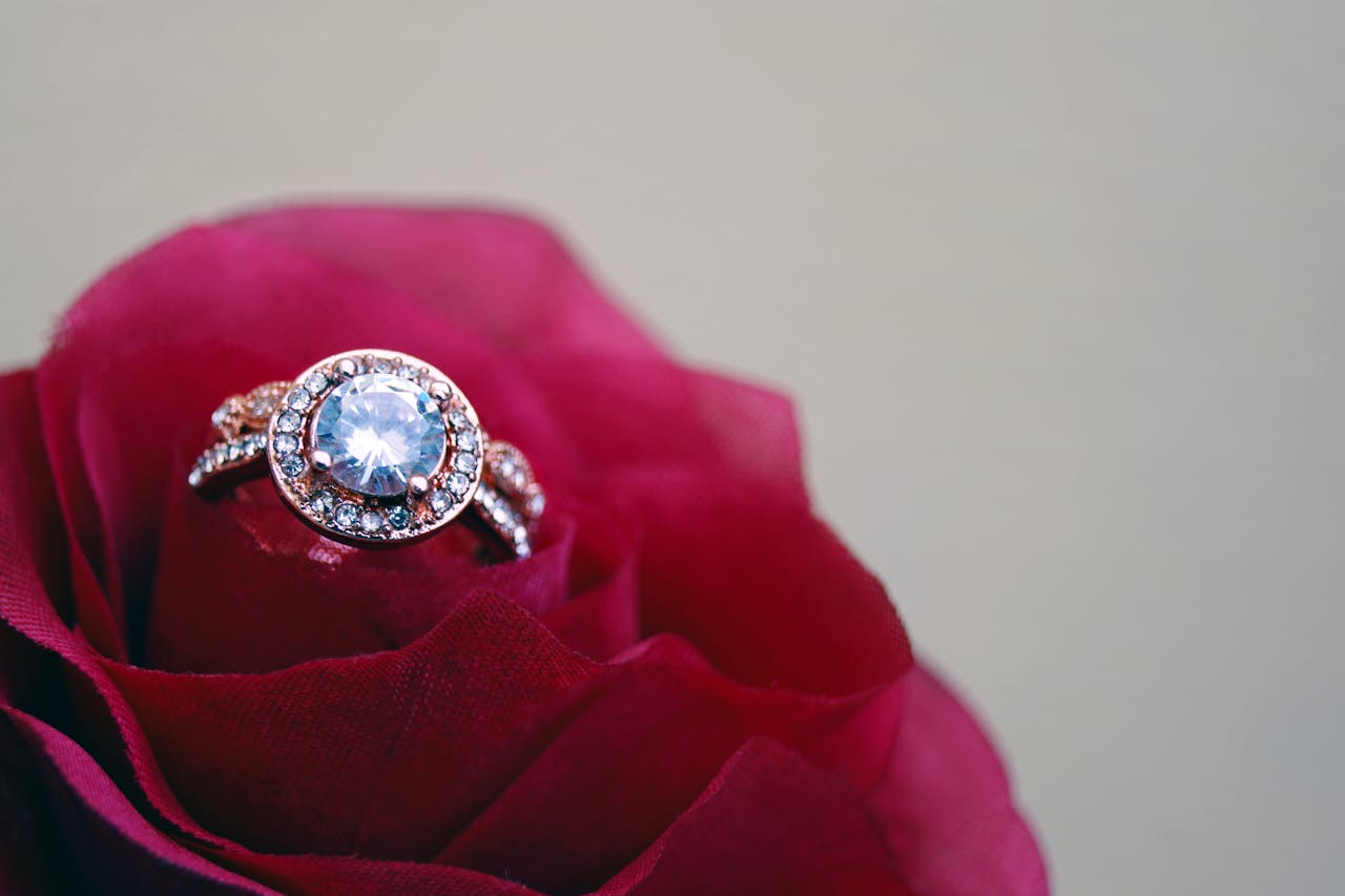 A close-up of a halo engagement ring with two rows of side stones displayed in the center of a red rose.
