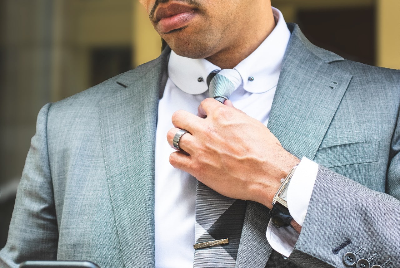 A close up of a man in a grey suit fixing his tie, showcasing silver wedding band