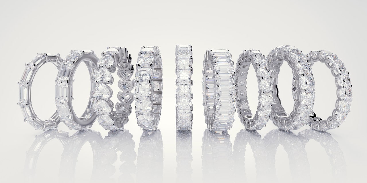 A row of elegant diamond eternity bands in various cuts and settings is displayed against a reflective white background.