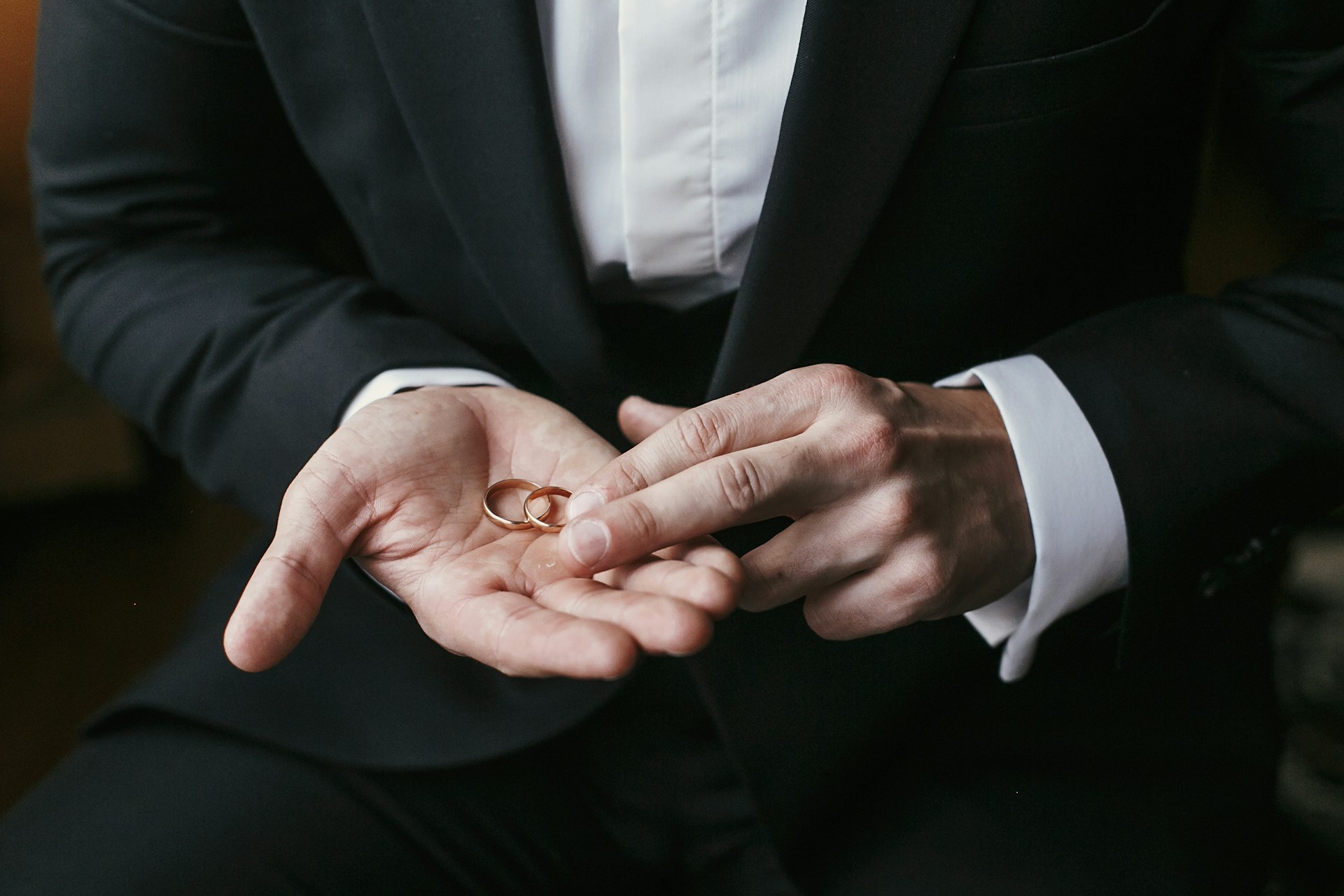 A close up of a man in a suit holding 2 thin yellow gold wedding bands.