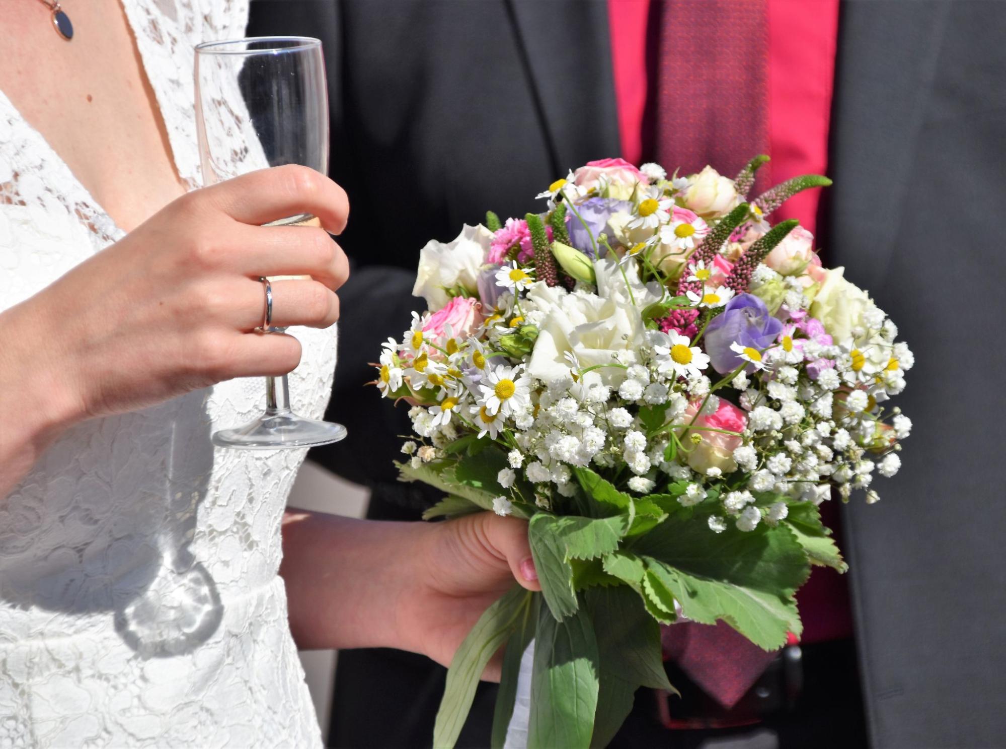 A bride holds a colorful bouquet and a glass of champagne, wearing a lace dress and wedding band, while a groom with a red shirt stands beside her.