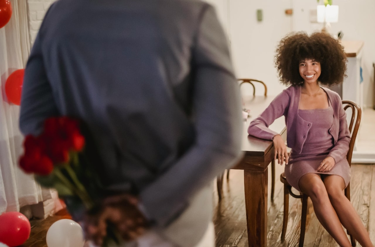 A smiling woman in a purple dress and cardigan sits at a wooden table while men hides a bouquet of red roses behind his back. A smiling woman in a purple dress and cardigan sits at a wooden table while men hides a bouquet of red roses behind his back.