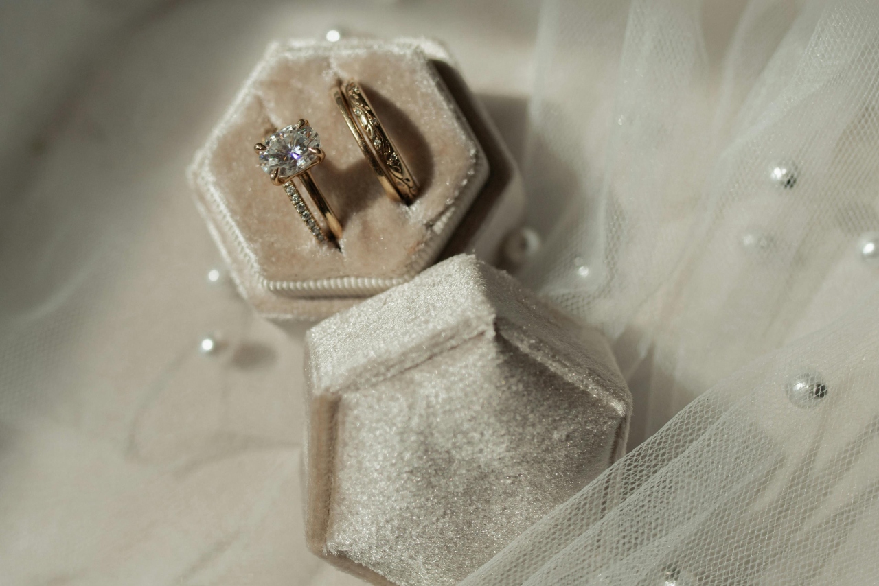 A yellow gold floral wedding band and a side stone yellow gold engagement ring with a radiant center stone in a hexagon ring box sitting on top of tulle with pearls
