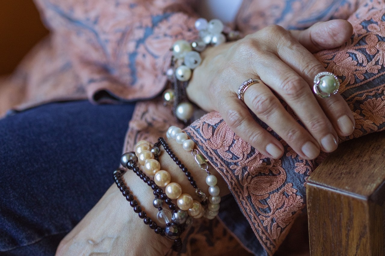 Close-up of a hand wearing layered pearl and beaded bracelets, a pearl ring, and wedding band, resting on a wooden armrest over a floral jacket.