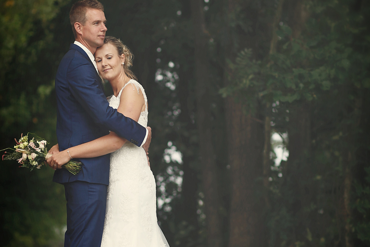 A couple embracing in the woods in their wedding attire, the woman’s head resting under the man’s head.