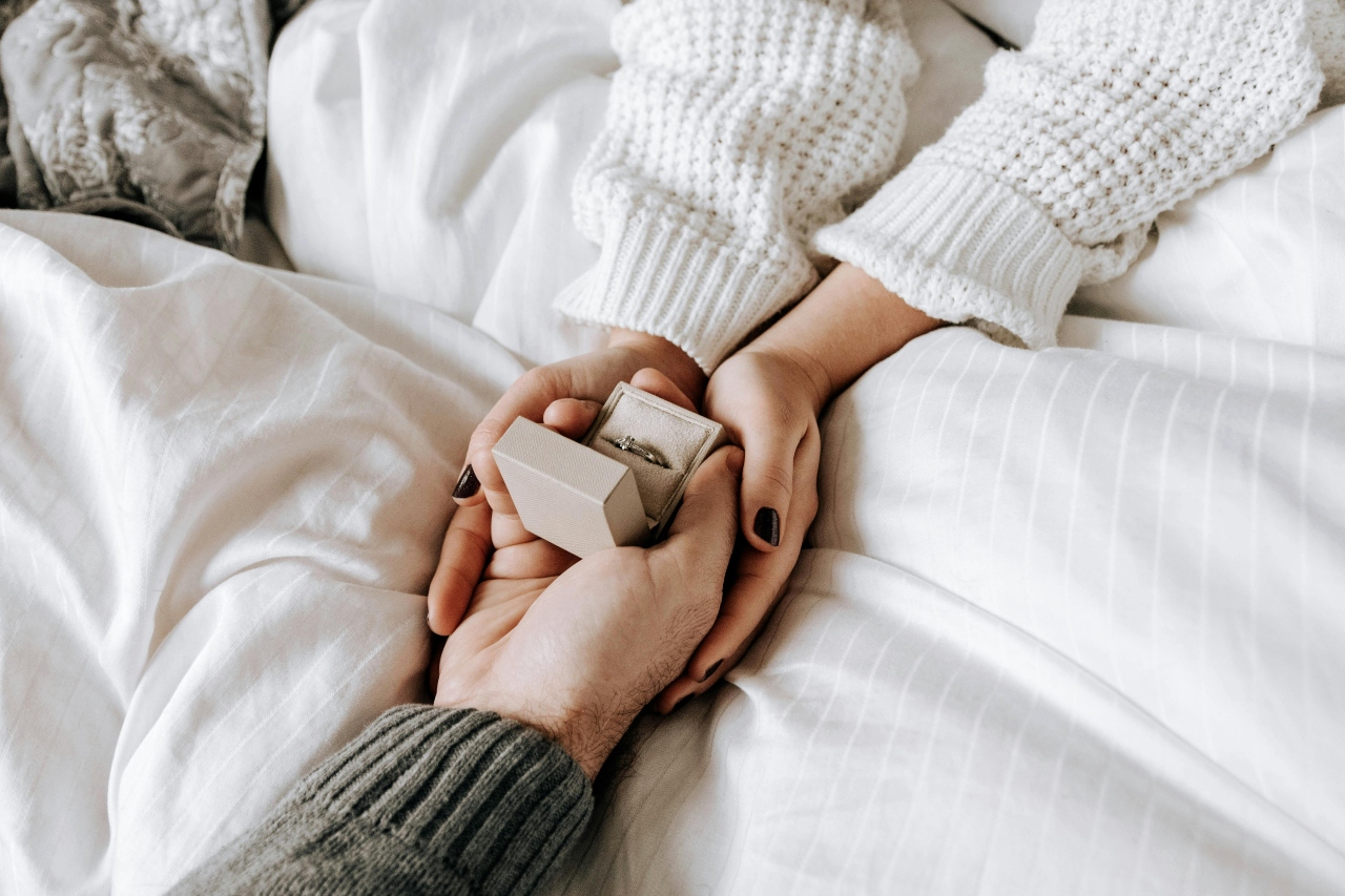 A comfortably dressed couple under a blanket, holding an engagement ring in their hands. A comfortably dressed couple under a blanket, holding an engagement ring in their hands.