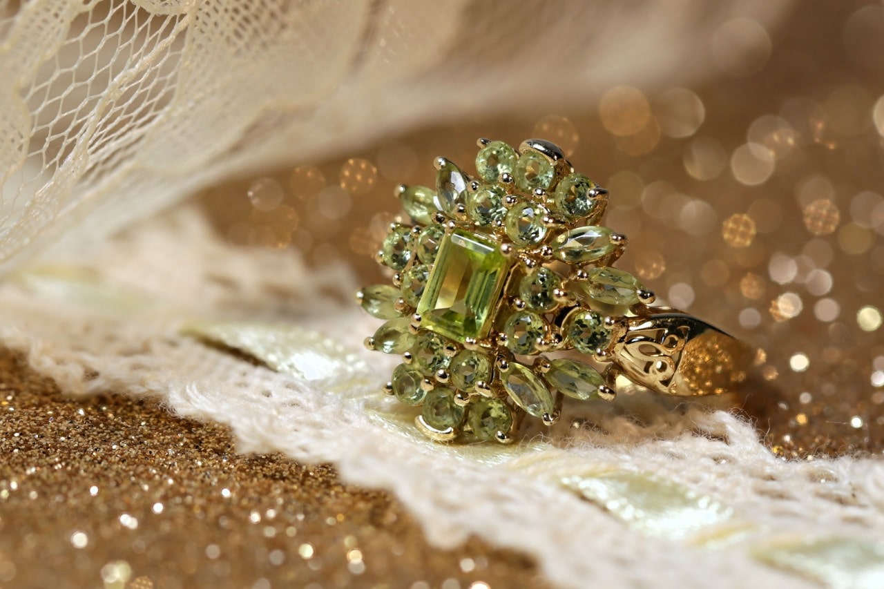 A yellow gold ring adorned with peridot gemstones rests on lace fabric with a sparkling, gold bokeh background. A yellow gold ring adorned with peridot gemstones rests on lace fabric with a sparkling, gold bokeh background.