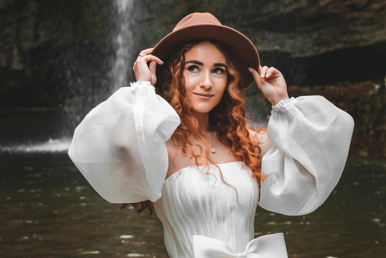 A young woman with curly hair in a white dress, wearing a diamond pendant and wide-brimmed hat stands by a waterfall. A young woman with curly hair in a white dress, wearing a diamond pendant and wide-brimmed hat stands by a waterfall.