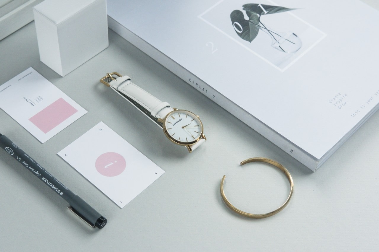 A close up of several items on white surface, including an open bangle, a wristwatch with white dial and strap,a grey pen and two small cards.